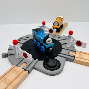 Könnte beinhalten: Ein blauer Spielzeugzug mit der Nummer 1 an der Seite steht auf einer schwarzen Drehscheibe. Ein gelber Spielzeugzug ist im Hintergrund. Das Set enthält graue Schienen, weiße Schranken mit roten Kreisen und rote Kugeln.