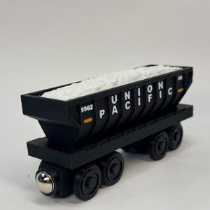 以下が含まれることがあります： 側面に白い文字でUNION PACIFICと書かれた黒いおもちゃの貨車。貨車の中には白い粒状の素材が入っており、側面には8562という数字が印刷されています。4つの黒い車輪が付いています。