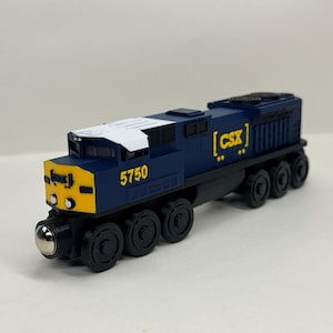 May include: A blue and yellow toy train engine featuring the number 5750 and the CSX logo printed on the side. The locomotive has a white roof section, black wheels, and a metallic front bumper detail, designed as a model railway vehicle.