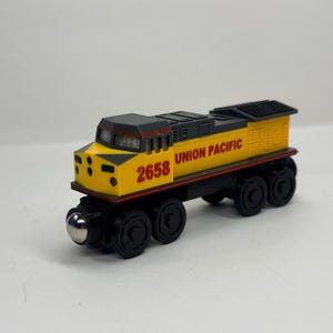 Puede incluir: Una locomotora de tren de juguete amarilla y negra con el texto UNION PACIFIC y el número 2658 en letras rojas. La locomotora tiene un techo gris oscuro, ruedas negras y una pieza de conexión frontal plateada sobre fondo blanco.