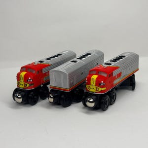 Puede incluir: Tres vagones de tren de juguete con un diseño en rojo y plata con detalles en amarillo. Dos locomotoras llevan la inscripción Santa Fe y los números 318 y 317, mientras que un vagón de pasajeros plateado se sitúa entre ellas. Tienen ruedas negras.