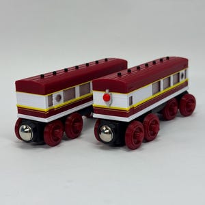 May include: Two toy train passenger carriages featuring a maroon and white colour scheme with yellow accent stripes. Each carriage has black roofs, multiple windows, and four red wheels with silver metallic hubs. One carriage displays a prominent red circular light.
