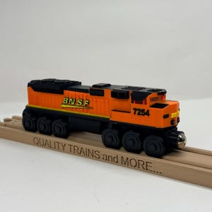 Könnte beinhalten: Eine orangefarbene und schwarze Spielzeugeisenbahn mit dem aufgedruckten Text BNSF und 7254 an der Seite. Der Zug steht auf einem hölzernen Gleisstück, das den eingravierten Text QUALITY TRAINS and MORE trägt.