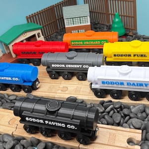 Puede incluir: Una colección de vagones de tren de juguete coloridos, cada uno con el texto "Sodor" y varios nombres de empresas. Los vagones son rojos, azules, naranjas, amarillos, grises, blancos y negros. Los vagones de tren de juguete están sobre una vía de madera.