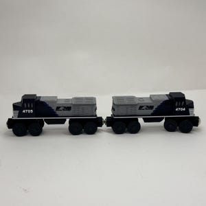 May include: Two toy train engines connected together, coloured black and grey. The left engine is numbered 4705 and the right engine is numbered 4704. Both feature the NS logo on the side, black wheels, and a detailed cab and engine compartment design.