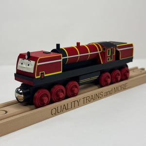Könnte beinhalten: Eine rot-schwarze Spielzeug-Dampflokomotive mit einem Cartoon-Gesicht an der Vorderseite, gelben Verzierungen und roten Rädern. Der Zug steht auf einem Holzschienenstück, in dessen Seite der Text QUALITY TRAINS and MORE eingraviert ist.