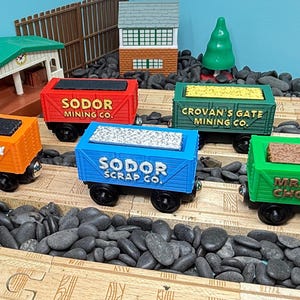 Puede incluir: Una colección de vagones de tren de juguete coloridos con varias etiquetas, incluyendo "Fred Pelhay Coal Co." y "Sodor Mining Co.". Los vagones son naranjas, rojos, verdes y azules, y están sobre una vía de madera con piedras negras. Un árbol verde y un pequeño edificio están en el fondo.