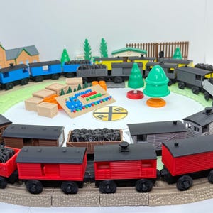 May include: A colorful toy train set arranged in a circle on a track. The set includes various train cars in red, blue, green, and orange, some loaded with coal. Additional elements include miniature trees, buildings, and a sign that reads "Quality Trains & Goods."