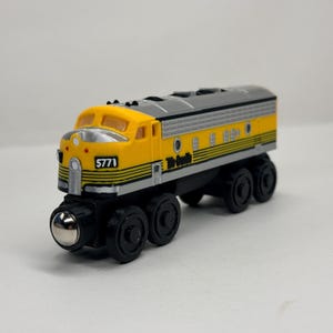 May include: A toy train engine featuring a yellow and silver body with black accents. The front displays the number 5771 in white on a black background. The side includes the text Rio Grande in black lettering. It has black wheels and a silver nose.