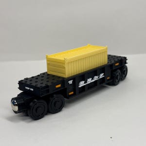 May include: A black toy flatcar train carriage featuring a yellow shipping container secured on top. The flatcar has black wheels, a metallic front connector, and white BNSF lettering printed on the side, with small orange rectangular accents.