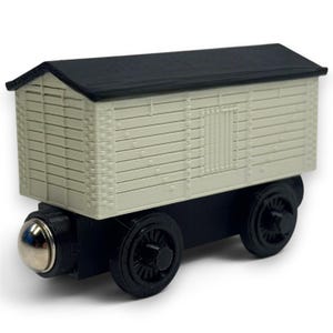 May include: A wooden toy train car with a black roof and wheels. The car's body is light beige with horizontal plank detailing. A silver-colored metal piece is on the front of the car.