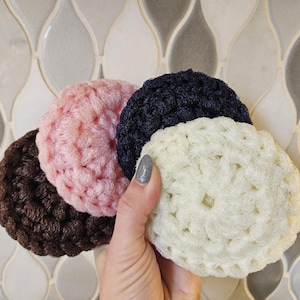 May include: Four crocheted circular items in brown, pink, navy blue, and cream are held in a hand. The items have a textured, looped design, suggesting they are handmade. The background is a decorative tile pattern.