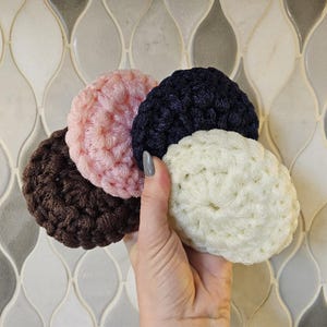 Puede incluir: Cuatro artículos redondos de ganchillo en tonos marrón, rosa, azul marino y crema. Los artículos se sostienen en una mano, mostrando la textura del hilo. El fondo presenta un patrón de baldosas decorativas.