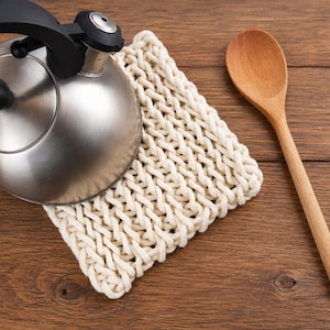 May include: A stainless steel tea kettle with a black handle and lid knob, resting on a square, off-white, thick-corded knit trivet, accompanied by a single light-colored wooden spoon positioned to the right of the trivet.