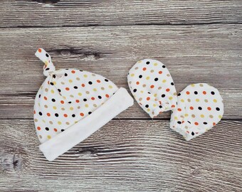 May include: A set of baby accessories in a white, yellow, orange, and black polka dot pattern. The set includes a hat with a knotted top and a pair of mittens.