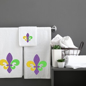 Puede incluir: Tres toallas de ba&ntilde;o blancas con dise&ntilde;os de lirios bordados en morado, verde y amarillo. Las toallas est&aacute;n colgadas en un toallero de cromo. Tambi&eacute;n hay toallas blancas en una cesta de alambre y en un estante.