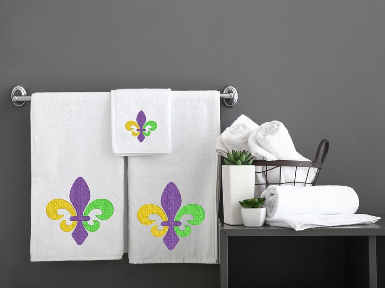 Puede incluir: Tres toallas de ba&ntilde;o blancas con dise&ntilde;os de lirios bordados en morado, verde y amarillo. Las toallas est&aacute;n colgadas en un toallero de cromo. Tambi&eacute;n hay toallas blancas en una cesta de alambre y en un estante.