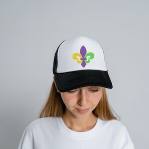 Puede incluir: Una gorra de camionero blanca y negra con un dise&ntilde;o de flor de lis morado, amarillo y verde en la parte delantera.