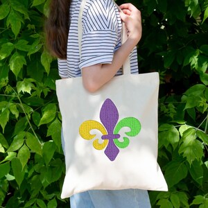 Puede incluir: Una bolsa de tela blanca con un dise&ntilde;o de flor de lis morado, verde y amarillo. La bolsa la lleva una persona que lleva una camisa a rayas.