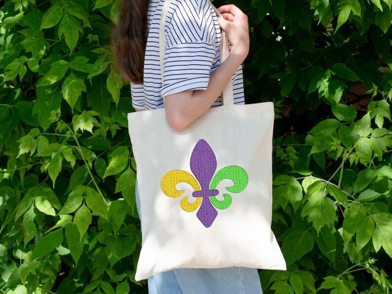 Puede incluir: Una bolsa de tela blanca con un dise&ntilde;o de flor de lis morado, verde y amarillo. La bolsa la lleva una persona que lleva una camisa a rayas.