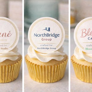 May include: Three cupcakes with white frosting and gold cupcake liners. Each cupcake has a round topper with a logo and text: "Luné Studio", "NorthBridge Group", and "Bloom Café". The text "crafted for" is also included.