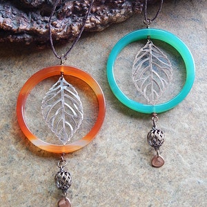 May include: Two necklaces with agate pendants featuring a silver leaf design. The pendants are attached to a brown cord and have a silver bead and a copper charm at the bottom.