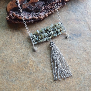 Puede incluir: Un collar de cadena de plata con un colgante rectangular hecho de piedras verdes y grises y una borla de plata.