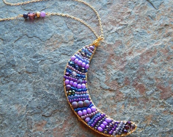 May include: A crescent moon pendant necklace with a gold chain. The pendant is made of wire wrapped with purple, blue, and silver beads.