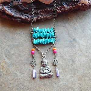 Puede incluir: Un collar de piedras turquesa con un colgante de Buda y cuentas rosas y naranjas. El collar está sobre una superficie de madera marrón.