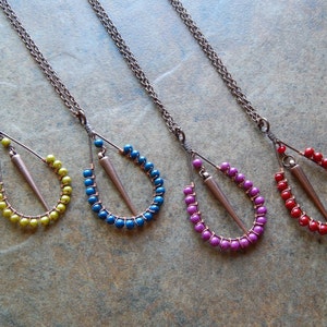 May include: Four necklaces with copper chain and a teardrop-shaped pendant made of copper wire and beads. The beads are in four different colors: green, blue, purple, and red.