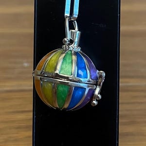 Könnte beinhalten: Halskette mit Anhänger in Form eines bunten Heißluftballons. Der Anhänger ist an einer rosa Kordel befestigt. Der Ballon hat einen silberfarbenen Metallrahmen und Verschluss.