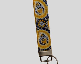 May include: A fabric wristlet key fob with a yellow and navy blue paisley pattern. The pattern includes yellow hearts and floral designs. The key fob has a silver-coloured metal clasp and key ring.