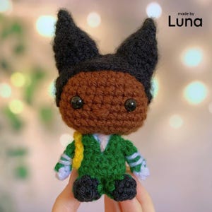May include: A small crochet doll with brown skin, black hair styled in two pointed ears, and black plastic eyes. The doll wears a green suit with white cuffs, black shoes, and a yellow accessory. The text "made by Luna" is visible in the top right corner.