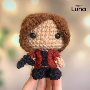 May include: A small crochet doll with brown hair, a beige face, and black eyes. The doll wears a red jacket over a black shirt and black pants. The text 'made by Luna' is visible in the top right corner of the image. The doll is crafted with yarn.