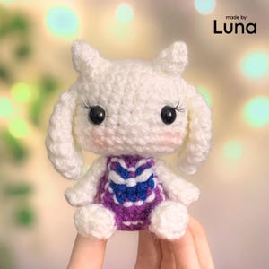 Könnte beinhalten: Ein weißes gehäkeltes Plüschtier mit langen Ohren, schwarzen Kunststoffaugen mit Wimpern und rosa Wangen. Die Figur trägt ein violettes Kleid mit einem weißen und blauen Blumenmuster. Der Text "made by Luna" steht in der oberen rechten Ecke.