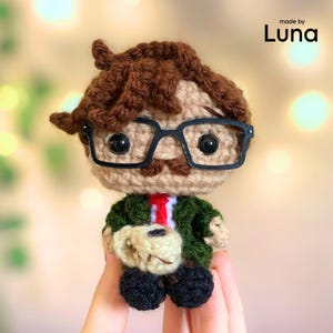 May include: A crochet doll with brown hair, black plastic glasses, and a mustache. The figure wears a dark green jacket over a white shirt with a red tie and holds a small beige object. The text "made by Luna" is printed in the upper right corner.