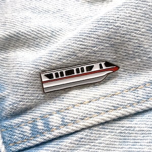 May include: A white and red enamel pin of a monorail train on a blue denim background.