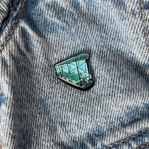 May include: A teal blue enamel pin of a vintage amusement park ride. The ride is a carousel with a glass roof and ornate details. The pin is a great addition to any vintage or amusement park lover's collection.