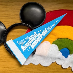 May include: A blue pennant with white text reading "There's a great big beautiful tomorrow, just a dream away" is displayed with a black Mickey Mouse ear headband and a felt rainbow. The pennant has a white border.