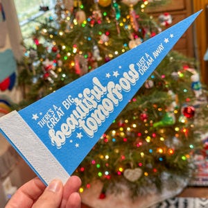 Puede incluir: Un banderín azul con texto blanco que dice "There's a big beautiful tomorrow, just a dream away." El banderín está colgado de un árbol de Navidad.