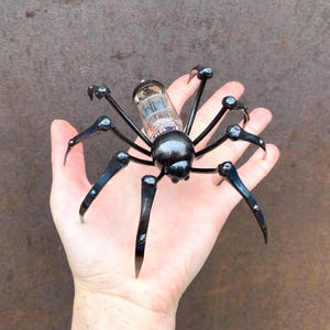 Op de afbeelding: Een handgemaakte metalen spinnensculptuur met een glazen buisvormig lichaam en zwarte poten. De poten van de spin zijn gebogen en eindigen in puntige punten. De sculptuur wordt in een hand gehouden.