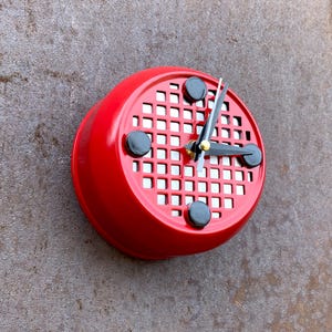 May include: A circular wall clock with a bright red frame and a silver-toned grid face. The clock features three black circular markers at the three, nine, and twelve o'clock positions, along with black hour, minute, and second hands at the center.