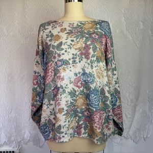 May include: A long-sleeved top with a boat neck, adorned with a floral print. The cream-coloured fabric is patterned with blue, pink, yellow, and green flowers and leaves. The material has a textured knit appearance.