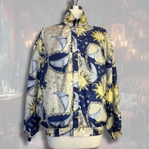 Puede incluir: Chaqueta vintage con cuello alto y cierre de cremallera. La chaqueta presenta un estampado repetido de veleros, soles y elementos celestes en azul marino, crema y amarillo. Tiene puños y cintura elásticos.