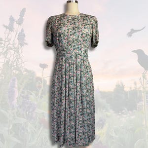 May include: A vintage floral dress displayed on a mannequin. The dress has a light blue base adorned with small pink, white, and green floral patterns. It features short sleeves and a pleated skirt, offering a classic style.
