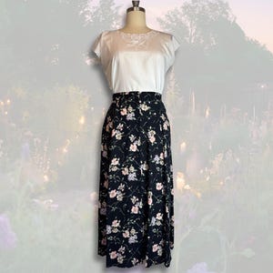 May include: A two-piece outfit on a mannequin. The top is white with a detailed neckline. The skirt is black with a floral pattern in pink, white, and green. The skirt is floor-length.