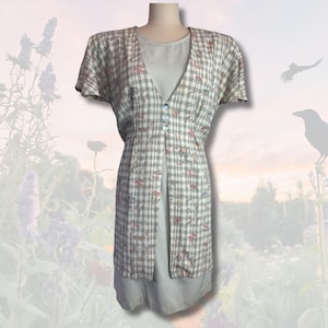 Puede incluir: Vestido vintage sobre un maniquí. El vestido presenta un estampado de cuadros grises claros y blancos con pequeños detalles florales rosas. Tiene una parte delantera abotonada y mangas cortas. El vestido está superpuesto a un enagua gris claro lisa.