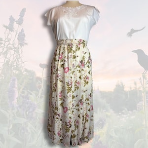 May include: A vintage dress with a white lace-trimmed bodice and short sleeves. The cream-coloured skirt has a floral pattern in pink and green. The dress has a high waist and a front slit. A classic design.