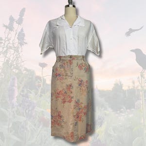 May include: A white short-sleeved button-down shirt paired with a beige floral print skirt. The skirt features a high waist and a knee-length hem. The floral pattern includes pink, red, and blue flowers.