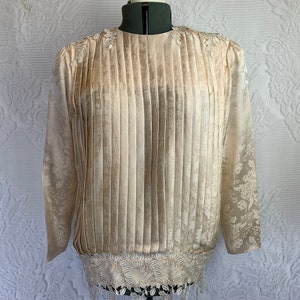 Könnte beinhalten: Eine cremefarbene, langärmelige Bluse mit einer plissierten Vorderseite und einer Spitzenborte. Die Bluse hat ein Blumenmuster und ist aus einem seidenartigen Stoff gefertigt.